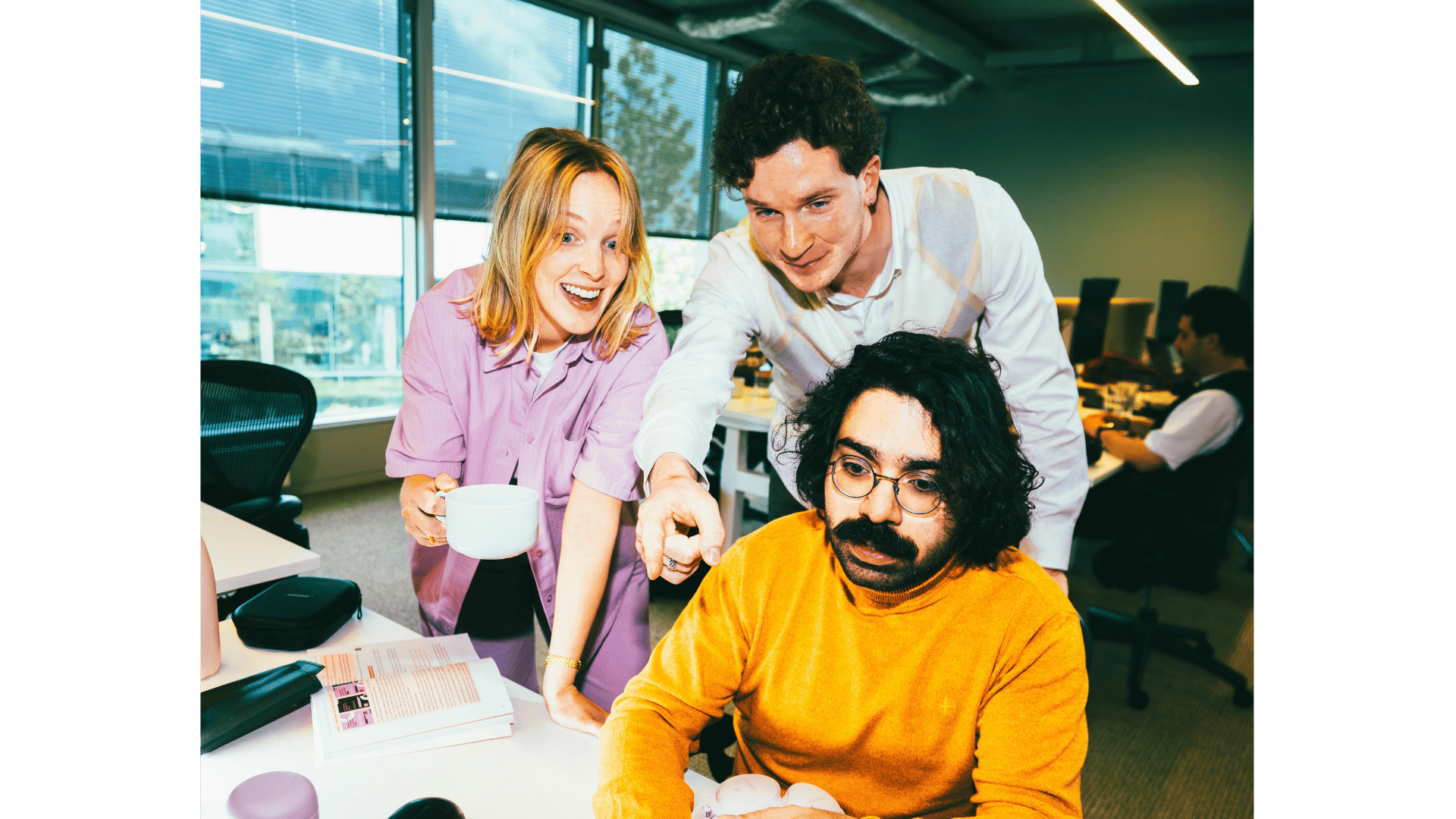 One young man sitting behind his desk with a young man and woman looking over his shoulders.